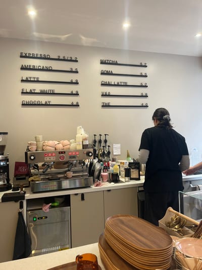 The image displays the interior of a coffee shop in Roubaix, France, centered on the service counter and a barista. A menu board composed of black text on a light grey wall presents two columns of items and their prices. The left column lists: "ESPRESSO 2.5/3.2", "AMERICANO 3.5", "LATTE 4.9", "FLAT WHITE 4.9", and "CHOCOLAT 4.8". The right column lists: "MATCHA 5.2", "GOMA 5.3", "CHAI LATTE 5.2", "V60 5.9", and "THE 4.0".

A person, viewed from behind, is positioned at the counter, wearing a black short-sleeved shirt over a light long-sleeved undershirt and dark pants. Their hair is secured with a patterned scrunchie. A partial view of a second individual's forearm and hand is visible on the far right.

The main work surface features a stainless steel La Marzocco espresso machine with three group heads, upon which multiple pink ceramic cups are stacked. A single small pink cup rests on the machine's drip tray. To the left of the espresso machine, a coffee grinder is visible. The counter behind the machine holds numerous syrup bottles with pumps, milk pitchers, and assorted coffee preparation tools and supplies, including a bag of coffee labeled "MU". Below the counter on the left, a stainless steel appliance, possibly an ice maker, is installed.

In the foreground, on a lower counter surface, a stack of approximately 15 oval, dark wood serving trays is prominently displayed. Adjacent to these trays, a container holds an assortment of spoons and other utensils, next to which are several small pink ceramic bowls. An amber-colored glass cup is partially visible in the bottom left corner. The ceiling is white with two recessed spotlights.
