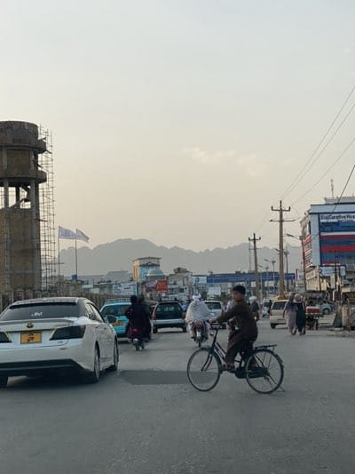 The image depicts a street scene in Kandahar, Afghanistan. In the foreground, a young man rides a bicycle. A white car is on the left. Various other vehicles, including motorcycles and cars, are visible in the traffic. Several pedestrians are walking along the street. In the background, mountains can be seen, and various buildings line the street, including a tall water tower structure with scaffolding. Power lines are also visible. The sky is overcast, and the light appears to be from late afternoon or early morning.