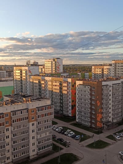 The image depicts a high-angle view of a residential area in Кондратово, Russia, under a sky with layered clouds during a sunset. Several apartment buildings with repetitive window patterns dominate the scene. Some buildings have colorful geometric patterns on their facades. Cars are parked in designated parking areas, and a couple of people are walking along a paved path. A grassy area is visible between the buildings. Overhead electrical wires are seen against the sky.