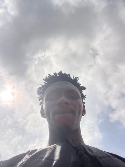 A low-angle, eye-level shot captures a person's head and upper torso against a bright, cloudy sky in Lokoja, Nigeria. The individual, with dark skin and dark, curly hair, is looking towards the camera with a subtle, serene expression. Their face is angled slightly upwards and to the left, with the right side of their face receiving more light. The sun, partially obscured by clouds, is visible in the bottom right, casting a bright flare and illuminating the edges of the person's hair and profile. The sky is predominantly filled with white and light grey clouds, indicating a partially overcast but bright daytime. The person is wearing dark-colored clothing, possibly a jacket or shirt, visible on the right side of the frame. No text is visible in the image.