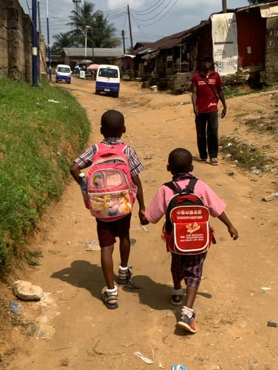 The image depicts a daytime scene on an unpaved, dusty street in Anantigha, Nigeria. In the foreground, two dark-skinned children, seen from behind, walk away from the viewer holding hands. The child on the left wears a plaid shirt, dark shorts, and sandals, carrying a pink backpack adorned with Disney Princess characters and a water bottle in its side pocket. The child on the right wears a pink shirt, plaid shorts, and sneakers, carrying a red and white backpack with Chinese characters and a phone number printed on it. Their shadows are visible on the ground. The dirt path shows scattered small pieces of debris.
To the left, a grassy bank borders a low wall. In the mid-ground, an adult dark-skinned male walks towards the viewer on the right side of the path, wearing a red striped polo shirt, dark trousers, and sandals, with a dark bag slung across his chest. Beyond the immediate path, the street is lined with rudimentary buildings constructed from various materials, many featuring corrugated metal roofs. Utility poles with overhead wires are visible. Further in the background, two white and blue commercial minibuses are present on the unpaved road, facing away from the camera. Palm trees and other vegetation are visible under a partly cloudy sky.