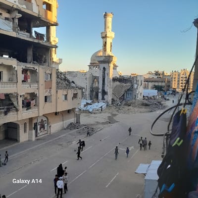 The image captures a street scene amidst war-torn buildings, with a mosque prominently standing partially destroyed. People walk on the street, suggesting movement and life persisting despite the surrounding devastation. The buildings on either side of the road are heavily damaged, exposing interiors and indicating the scale of destruction. The presence of a clear sky contrasts sharply with the ground-level chaos, creating a poignant juxtaposition.