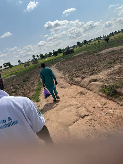 The image shows a person in a teal-colored outfit walking across a dirt path with a purple object in their hand. They are viewed from behind. To the left of the path is a grassy field with scattered trees and some small buildings, suggesting a rural or semi-rural setting. In the background, there are clouds in a bright sky, indicating it is daytime and likely fair weather.
In the foreground, another person's arm wearing a white long-sleeved shirt is visible. The shirt has some text printed on it, though it is partially obscured by a blur. Visible text reads "ain A mentation". The overall scene appears to be outdoors in a warm climate. The composition and the orientation of the image suggest it might have been taken while the photographer was also in motion or from a low vantage point. The location context provided is Jalingo, Nigeria.