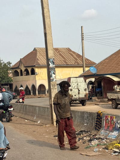 The image depicts a daytime street scene in Lafia, Nigeria, characterized by urban infrastructure and public activity.
In the foreground, a male individual stands facing slightly right, looking downward. He has dreadlocks and a full beard, appearing unkempt. He wears a dark, soiled short-sleeved t-shirt with obscured text, possibly reading "SASTODAY SAI JOS," and dirty, reddish-brown pants. His feet are bare or covered by minimal, worn footwear. To his left, a thick, grey concrete utility pole stands, featuring two political posters with black-and-white images of a man's head, one labeled "M. ENITO" and the other "M. MAGA."
The central figure is positioned on a dirt shoulder beside a paved road. A low concrete barrier separates this shoulder from the main roadway. Along the base of this barrier, to the right of the man, an accumulation of discarded refuse, including plastic, paper, and other debris, is visible. Two flattened posters, one displaying "STAR RADLER" branding, and a piece of striped fabric (blue and yellow/white) are also on the ground near the trash.
On the paved road to the left, two individuals on motorcycles are present. One rider wears a light blue shirt and blue jeans, while the other, wearing a red shirt and white cap, is seated on a dark motorcycle, facing away.
In the mid-ground, behind the concrete barrier and the central figure, a white, dilapidated panel van with visible rust and dirt is parked on a dirt patch. To the right of the van, a small, open-front establishment with a brown corrugated metal roof and a blue satellite dish is visible. A person is indistinctly seated inside this structure. Further right, a thinner utility pole with multiple power lines extends across the frame.
The background is dominated by a two-story building with yellow-painted walls and a multi-gabled brown corrugated metal roof. The ground floor features a series of arched openings. The sky above is light blue with a single small white cloud. Green foliage from trees is visible on the far left.