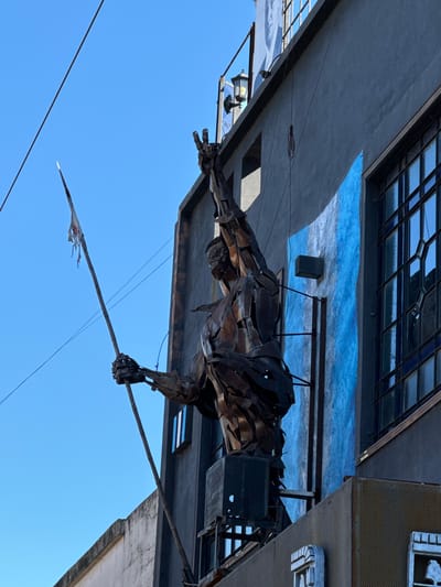 The photo depicts a sculpture of a man holding a spear erected on top of a building in Buenos Aires, Argentina. The sculpture is constructed from metal sheets welded together. The man is in a lunge position, poised to throw the spear. The building has windows and several horizontal blue and white stripes painted beneath it. The sky is clear blue.