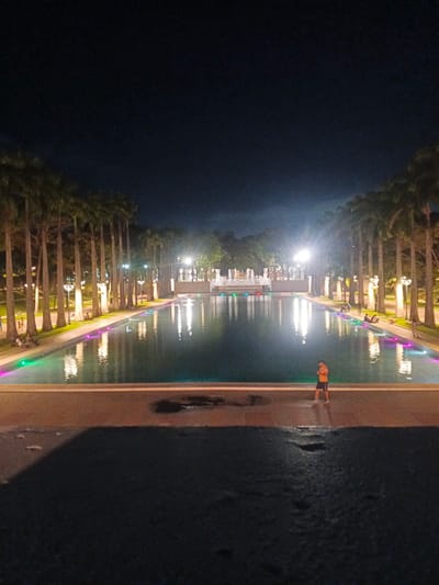 The image captures a nighttime view of the Parque del Este in Caracas, Venezuela, featuring a large, rectangular reflecting pool. The pool is illuminated from beneath with colorful lights, casting vibrant reflections on its surface. Tall palm trees line the perimeter of the pool, creating a distinct avenue. Several individuals are present in the scene. In the foreground, a lone person stands on the edge of a paved area overlooking the pool. Further back, along the pool's edge and beneath the palm trees, other people are seen sitting or walking, some interacting in small groups. In the background, structures and more lighting are visible, suggesting the park extends beyond the immediate scene. The overall atmosphere is serene and illuminated, highlighting the park's features at night.