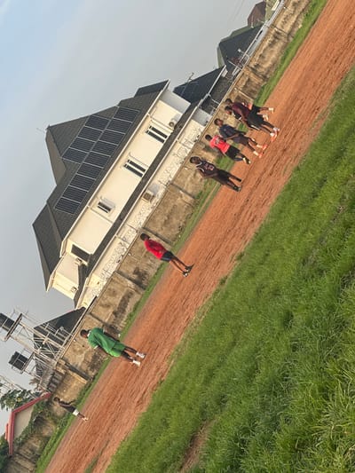 The image shows a group of young men in athletic wear standing on a dirt track next to a grassy area, with a modern white house featuring solar panels in the background. The scene appears to be outdoors during the day, with clear skies suggesting fair weather. Some of the men are standing in a line, while others are more spread out. One person in a red shirt is captured mid-movement, possibly engaged in an athletic exercise. Another individual in a green shirt and shorts is standing further down the track.
The architectural style of the house, with its clean lines and prominent solar panels, hints at a modern residential setting. The dirt track and grassy area suggest a sports field or training ground. The overall atmosphere seems relaxed and focused on physical activity.
No specific time of day is definitively indicated beyond daylight, but the soft lighting might suggest early morning or late afternoon. The presence of the house and the general upkeep of the area suggest a populated or developed location. The people's attire and the setting imply an informal sports or training session. The image is taken at an angle, with the horizon line tilted.
No visible text is discernible in the image. The context provided states the location is Abuja, Nigeria, which aligns with the visual cues of modern architecture and an outdoor environment.