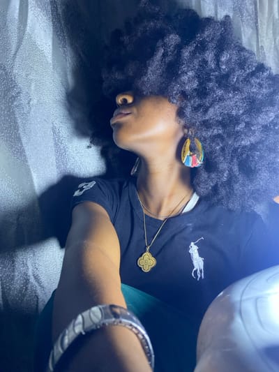 An individual with dark skin and voluminous, dark, curly hair, which obscures the upper face, is depicted in a medium shot. The subject is seen in profile, looking upwards and to the right of the frame, with the left arm extended towards the camera. They wear a black short-sleeved t-shirt featuring a white Polo Ralph Lauren logo on the chest and a white number '3' on the left sleeve. Two necklaces are present: one thin silver-colored chain and one thicker gold-colored chain with a quatrefoil-shaped pendant marked '5'. A large, multi-colored (yellow, red, blue, green) teardrop-shaped earring is visible on the right ear. A wide, silver-colored bracelet with an oval or striped design adorns the extended left forearm. A green garment is partially visible beneath the t-shirt. The background consists of a light grey, textured material, possibly a curtain or upholstery. A bright, white, curved object, likely an artificial light source, occupies the lower right foreground. The scene is illuminated by a strong light source from the bottom right, creating shadows on the left side of the face and behind the head. The specific location in the city of Iranla, Nigeria, cannot be determined from the visual information.