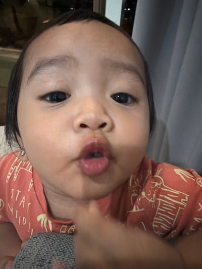 A close-up, eye-level, portrait shot shows a young East Asian child with dark hair, dark eyes, and full lips puckered as if to give a kiss. The child wears a coral-colored t-shirt with a white line-drawn animal print and the text "STAY WILD!!" The child is on a gray, woven surface, possibly a blanket or cushion. The background is blurred, showing a light gray curtain to the right and a darker, out-of-focus area to the left, with hints of lights. The image appears to have been taken indoors. The child's facial expression and pose suggest playfulness or an attempt to interact with the camera. The lighting is warm and diffused, casting soft shadows on the child's face.