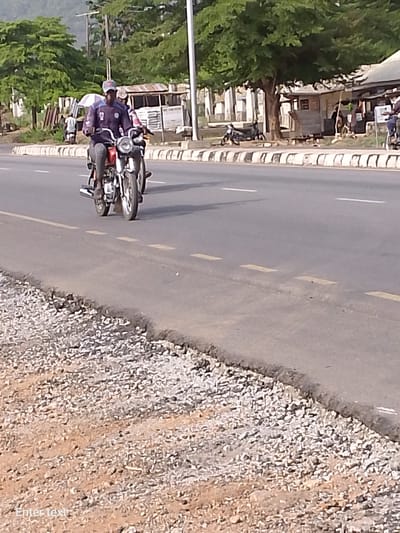 A medium shot depicts two people on a red motorcycle traveling on a paved road in Adankolo, Nigeria. The road features intermittent yellow lines and a concrete curb separates it from a gravelly area on the right side. On the opposite side of the road, there are several small buildings and trees.