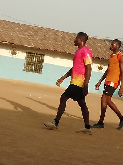 The scene is set outdoors in Akwanga, Nasarawa State, Nigeria, identifiable by the Nasarawa State emblem visible on a building facade. The ground consists of reddish-brown earth, exhibiting long shadows, indicative of either early morning or late afternoon light. The sky is clear.
Two adult males are walking from left to right across the frame. The male in the foreground wears a short-sleeved t-shirt with a pink-to-yellow gradient, black athletic shorts, and light-colored athletic shoes. Perspiration is visible on his face and neck. The second male, positioned behind and to the right, wears an orange sleeveless athletic vest over a white and orange garment, black athletic shorts with a white stripe and the number "5" on the left leg, and dark-colored shoes. He also exhibits visible perspiration. Both individuals are moving in the same direction, appearing to engage in physical activity.
In the background, a single-story building features a weathered, corrugated metal roof. The building's walls are painted light blue on the upper section and white on the lower section. A window secured with white metal bars is visible in the light blue section of the wall. The circular emblem of Nasarawa State is affixed to the building's light blue facade, both left of the window and to the right of the foreground male.