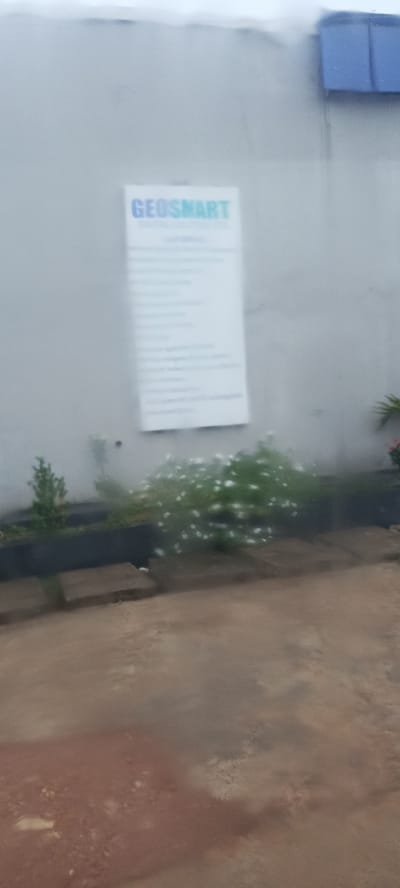 The image shows a sign on a wall with the text "GEOSMART DIGITAL SOLUTIONS". Below "GEOSMART" there are several lines of text, but they are not clearly legible. The sign is white with blue and green lettering. To the left of the sign, there is a small green bush. In front of the wall, there is a bed of white flowers. The ground in the foreground appears to be dirt or gravel. The wall is light gray. There is a blue structure visible on the top right corner of the image. The image appears to have been taken outdoors during the daytime. The weather appears overcast or possibly rainy, as indicated by the slightly blurred and hazy appearance of the image. The overall scene suggests an exterior view of a building, likely a business premises, in Bukuru, Nigeria.