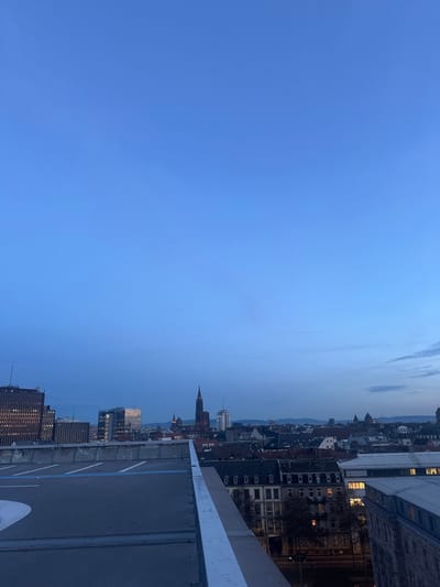 The image presents a panoramic view of Strasbourg, France, observed from a high vantage point, likely a rooftop or helipad, during dusk or early evening. In the immediate foreground, a dark grey, flat surface resembling a helipad is visible, marked with white and blue lines, and bordered by a white railing.

The midground showcases the city's extensive skyline, featuring a blend of traditional European architecture with dark-tiled roofs and lighter facades, interspersed with some modern buildings. A distinctive, reddish-brown stone spire, characteristic of a large cathedral, dominates the mid-left section of the cityscape, strongly resembling the Strasbourg Cathedral. Lights are illuminated in several building windows, indicating the twilight hour.

The background is dominated by a vast, clear blue sky that gradually lightens towards the horizon, where the faint outlines of distant hills or mountains are visible. The overall scene conveys a serene atmosphere as the city transitions from day to night. There are no people visible, and no specific activities are taking place. No discernible text is present other than the abstract markings on the foreground surface.