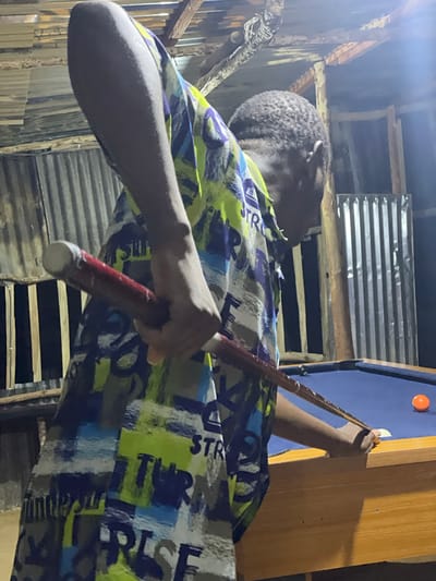 A young Black male stands at a blue billiard table, his body angled as he prepares to strike a white cue ball with a red cue stick. He is wearing a short-sleeved shirt with a colorful abstract pattern of green, blue, white, and purple brush strokes and text. The billiard table has a wooden frame and a bright orange ball rests in the corner pocket. The background consists of corrugated metal walls and a rustic wooden ceiling with exposed beams, suggesting an outdoor or semi-outdoor establishment, potentially in Wumat, Nigeria. The lighting is dim, with a bright light source illuminating the upper right portion of the scene.