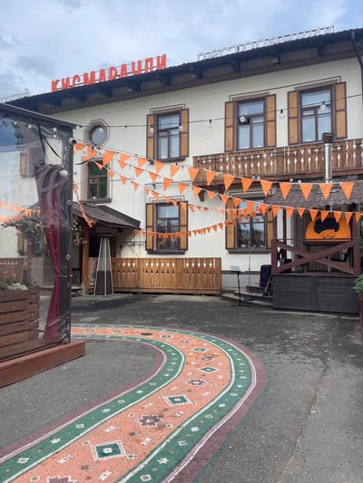 The image shows the exterior of a building in Perm, Russia. The building has a cream-colored facade with brown wooden window frames and shutters. Orange pennant banners are strung across the front of the building. A large sign with red Cyrillic letters is mounted on the roof. A portion of a painted rug pattern is visible on the pavement. The sky is overcast.