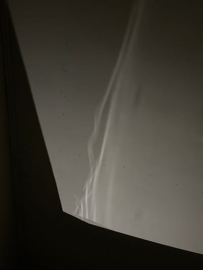 The image shows a dark, abstract composition with a prominent diagonal shadow on the left, dividing the frame. The right side is a lighter, off-white surface with subtle, undulating streaks of light that appear to be reflections or refractions. A few small, dark specks are scattered across the lighter surface, possibly dust or imperfections. The overall impression is one of subdued light and shadow play on a textured surface. No specific location within Lhokseumawe, Indonesia, can be determined from the visual information provided.