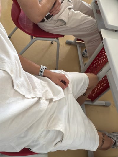The image depicts an indoor scene, likely a classroom or lecture hall, with two individuals seated at desks. The floor is a light brown linoleum. In the foreground, a person wearing a white linen shirt and white shorts is seated on a red chair. The person has a white and black patterned watch on their left wrist. A tattoo is visible on their right thigh, with the visible portion reading "GOES AM."

In the background, another person is seated on a similar red chair, wearing light-colored pants and a dark belt. Part of a white desk with a red perforated metal support is visible to the right. The lighting suggests an artificial light source, likely overhead. The image does not contain any specific visual cues that would allow for its location to be identified as Plovdiv, Bulgaria.