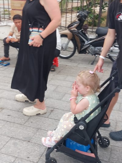 The image depicts an outdoor scene with several people and objects on a paved walkway, likely in Nha Trang, Vietnam. In the foreground, a person wearing a long black dress and white slide sandals is partially visible, with their hand resting on a small, colorful striped bag. To the left, a person wearing a black t-shirt is also visible, with an arm outstretched towards a stroller.
The central focus is a young child in a stroller, wearing a light green dress with a daisy pattern. The child has blonde hair and is touching their face, appearing distressed. A pink hair clip adorns their hair, and a pink bracelet is on their wrist. The stroller itself is black with some blue fabric visible inside.
Behind the stroller, a motorcycle is parked. Further back, a young man in a light-colored shirt and dark pants is visible, also appearing to be looking at something or someone. The ground is made of grey paving stones. The lighting suggests it is daytime. The overall atmosphere appears to be a casual outing, possibly in a public area.