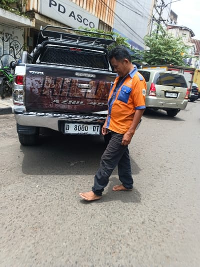 A barefoot man in an orange and blue uniform and dark pants walks on asphalt beside a Toyota Hilux pickup truck. The truck has a black rack above the cab, and “HILUX AZRIL 484” is painted on the tailgate in a faded brown font. A gold minivan is parked behind the truck. The background includes a building with “PD ASCO” on it and overhead wires.