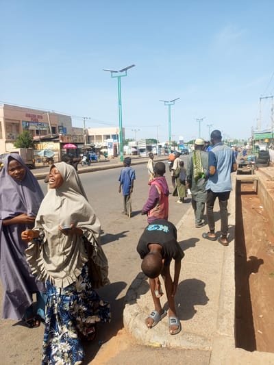The image depicts a street scene in Tempure, Nigeria, likely during daytime given the bright sunlight casting distinct shadows. The setting is a paved road with buildings and shops lining one side. Several individuals, including men, women, and children, are present on the street and sidewalk.
In the foreground, two women wearing hijabs are engaged in conversation. One woman, on the left, wears a dark purple hijab. The woman on the right, who is laughing or speaking animatedly, wears a lighter grey hijab and a colorful floral patterned skirt. She is holding a mobile phone. To the right of the women, a young boy in a black t-shirt and grey sandals is bent over, appearing to be picking something up from the ground or tying his shoe. Further down the street, other individuals are visible walking, talking, or standing. A man in a blue shirt and dark pants walks away from the camera, while other people are seen in the background near shops and along the road.
The environment suggests a typical urban or semi-urban street with ongoing daily activities. Streetlights with solar panels are visible along the road, indicating infrastructure development. Shops with signs are present, though the text on most is not clearly legible. One sign reads "ALHAMDU LILLA" and another appears to be "HAWEE'S". The overall atmosphere seems casual and bustling, with people going about their day under a clear, bright sky.