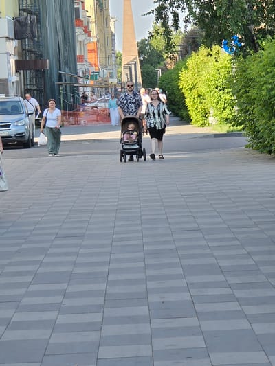 This media file depicts an outdoor scene in Krasnoyarsk, Russia, on a sunny day. A group of people are walking on a sidewalk paved with light gray rectangular tiles. The foreground features a person in a white t-shirt and green pants, and another person in a striped shirt and dark pants. A blue car is parked to the right.
Further back, a family appears to be out for a stroll. A woman wearing a black and white patterned top and black shorts is pushing a baby stroller. A man with sunglasses and a dark patterned shirt is walking beside her. A baby can be seen in the stroller. Other individuals are visible in the background, some walking and others standing.
Behind the people, there are trees with lush green foliage on the left side of the frame. The background shows buildings with colorful facades, suggesting an urban environment. Construction fencing with orange mesh is also visible, indicating ongoing work. The lighting suggests it is daytime, likely late morning or afternoon. The general atmosphere appears relaxed and casual, consistent with a leisurely outing on a pleasant day. No visible text is present in the image.