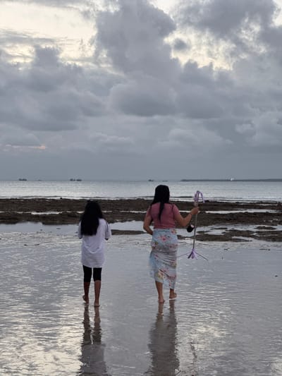 The image depicts two young females on a wet, sandy beach, with the ocean in the background. One girl, in the foreground, wears a white t-shirt and black shorts, standing with her feet in shallow water, her reflection visible on the wet sand. Behind her, another girl is partially visible, wearing a pink t-shirt and a floral skirt, holding a purple object on a stick.
The scene is set at what appears to be low tide, with dark, rocky formations jutting out from the water. The sky is overcast with dramatic, grey clouds, suggesting a storm might be approaching or has recently passed. The lighting is dim, indicating it is likely late afternoon or early evening. The ocean stretches out to the horizon, where a few distant boats or ships are visible.
The overall mood is calm and serene, despite the cloudy weather. The reflections on the wet sand add a reflective and slightly surreal quality to the image. The location, Kuta Selatan, Indonesia, is known for its beaches, and this scene captures a quiet moment by the sea. No visible text is present in the image.