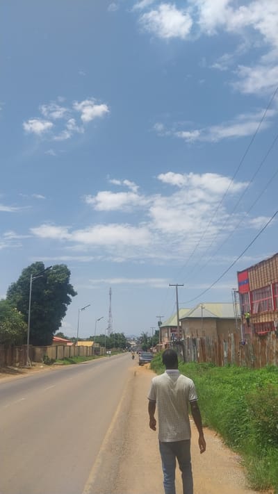 The image depicts a sunny day with a clear blue sky dotted with scattered white clouds. A man, seen from behind, is standing on the side of a paved road. He is wearing a patterned short-sleeved shirt and blue jeans. The road stretches into the distance, with a few vehicles visible, including a bus and motorcycles.
On the left side of the road, there are buildings with corrugated iron roofs and some construction materials. Further down the road, there are power lines and a telecommunications tower. Lush green grass and trees line the right side of the road, suggesting a semi-rural or suburban setting in Bukuru, Nigeria. The sun is high in the sky, indicating it is likely midday. The overall impression is one of a typical day in a developing area, with the man walking or standing near the road. No text is clearly legible in the image.