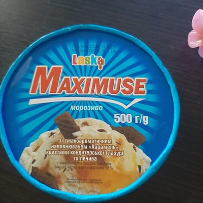 The image is an overhead shot of a blue ice cream container with the brand "Laska" and the product name "MAXIMUSE" prominently displayed in large orange letters. The container features a sunburst design emanating from the center. Text in Ukrainian is visible, including "морозиво" (ice cream) and "500 г/g" indicating the weight. Below this, there is a description of the ice cream's flavor and ingredients, and a note about the serving suggestion being of a fantasy character. The ice cream itself is visible within the container, appearing to be vanilla-flavored with chocolate pieces and wafer. To the right of the container, a portion of a pink, multi-petaled decorative flower is visible. The background is a dark, textured surface, likely a table. There is no indication of the city of Sambir, Ukraine, or any people in the image.