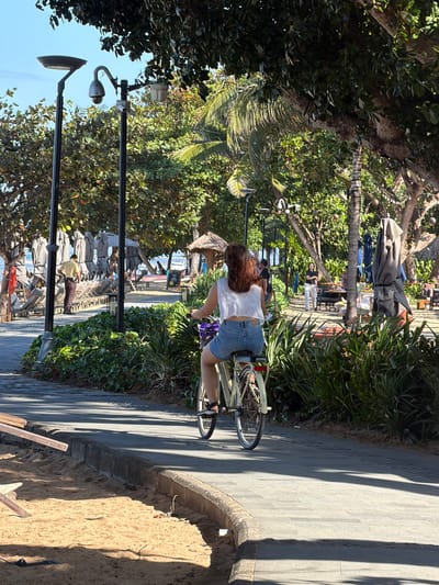 The image depicts a sunny day along a paved walkway adjacent to a sandy beach in Denpasar, Indonesia. In the foreground, a person is riding a cream-colored bicycle, viewed from behind. The cyclist is wearing a white, cropped sleeveless top and denim shorts, with their hair styled in loose waves. The bicycle has a purple basket. To the left of the walkway, there is a sandy area with some wooden debris.

The walkway is bordered by lush green foliage. In the mid-ground, several tall black lampposts with security cameras are visible. Beyond the lampposts, there is a resort-like area with rows of folded beach chairs and umbrellas, and thatched-roof structures. A few people are scattered throughout this area, some walking and others seated. The background reveals the ocean with boats in the distance and a clear blue sky. The overall scene suggests a recreational area, likely a beach promenade in a tropical setting.