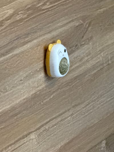 This is a close-up, high-angle shot of a small, cartoonish object with a white, rounded body and yellow fins or feet. The object has black dot eyes and a crescent-shaped mouth drawn on its face. The top of the object is a concave depression filled with a green, granular substance, likely catnip or a similar dried herb. The object is resting on a wooden surface with a visible grain pattern, appearing to be a floor. The lighting suggests an indoor setting, possibly during the daytime. There is no visible text in the image. The scene is simple and static, focusing on the small object and its texture against the wood.