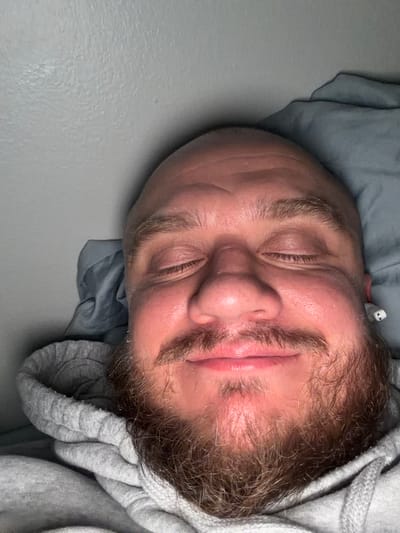 A close-up, upside-down photograph depicts a man with fair skin, a bald head, a full brown beard, and closed eyes, suggesting sleep. He is lying on what appears to be a grey bedsheet, with part of a grey hooded sweatshirt visible around his face. An electronic device, possibly an earbud, is seen near his right ear. The background is a plain light grey wall. The location is unknown, but given the query's inclusion of Morton Grove, United States of America, it is presumed the photo was taken there, although there is no visible evidence in the image to confirm it.