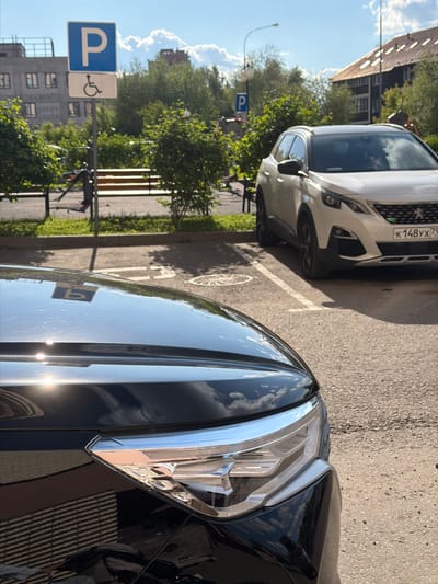 The image presents a daytime outdoor scene depicting a parking area in Odintsovo, Moscow Region, Russia. The foreground features the front section of a dark, reflective vehicle, specifically its glossy black hood and the right headlight assembly. Strong sunlight reflects off the hood, creating bright glares and mirroring the sky, clouds, and an inverted "P" parking sign. The headlight unit displays internal LED elements and a chrome trim.

In the midground, a white Peugeot 3008 SUV is parked, facing away from the viewer. Its front grille, headlights, and part of the bumper are visible. A Russian license plate, "K 148 YX 79," is affixed to its front, with the "79" regional code indicating the Moscow region. The asphalt parking surface beneath the vehicles features a faded white parking space line and a circular metal manhole cover to the left of the white vehicle.

Adjacent to the parking area, a blue and white parking sign is mounted on a metal pole. The sign displays a capital "P" above a white rectangular panel featuring the international symbol for accessibility (a wheelchair user). Another "P" parking sign is partially visible further back. Behind the signs and parking area, a strip of green grass, several bushes, and trees are present, along with a brown wooden bench and a partially visible metal outdoor exercise apparatus.

The background includes multiple buildings under a bright blue sky with scattered white cumulus clouds. On the left, a multi-story, possibly concrete-colored building with numerous rectangular windows is partially visible. On the right, a darker building with a distinct brown, pitched roof and multiple roof vents is prominent. A streetlight pole extends into the upper right corner of the frame. No human figures are present. The lighting indicates strong daylight with sharp shadows.