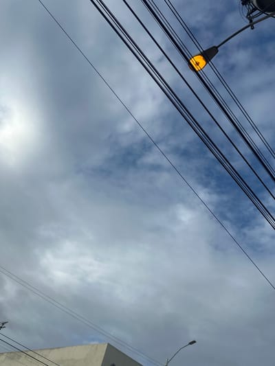 An outdoor, low-angle shot captures a cloudy sky with power lines crisscrossing the frame. A street lamp with an illuminated orange bulb is prominently featured on the right side, positioned above the power lines. In the lower left corner, the top corner of a light-colored building is visible. The scene appears to be in Porlamar, Venezuela, evidenced by a streetlamp design common in the city, indicating a daytime or twilight setting given the lamp's illumination against the ambient light.