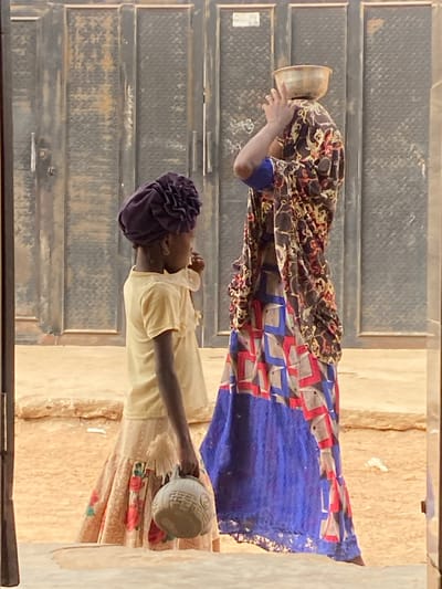 Two women stand in front of a gray corrugated metal wall. The woman above has a patterned dress, holding a pot to her face. The lower woman wears a light yellow shirt and a purple head covering, extending her arm with a patterned vessel. Both individuals appear to be outside, with a sandy, textured background visible.