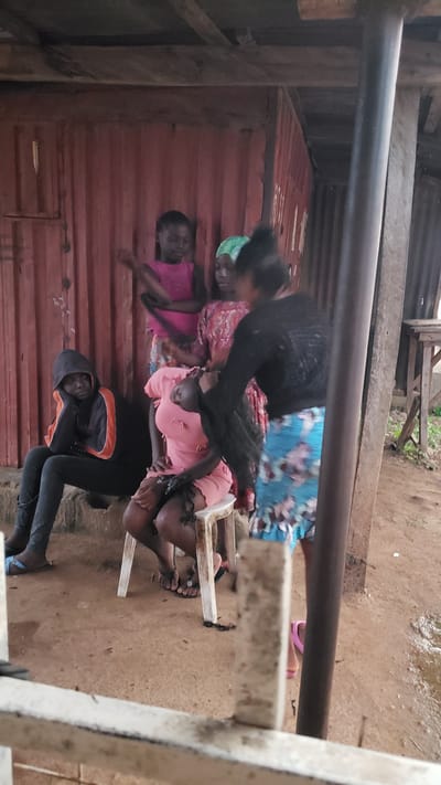 A group of young women and girls are gathered in an outdoor, makeshift shelter in Narayi, Nigeria. The structure appears to be made of corrugated metal walls and a wooden roof. In the foreground, a young woman with dark, long hair is seated on a white plastic stool. Another young woman, wearing a pink dress, is seated next to her, also on the stool, with her head bowed. A third young woman, wearing a black hoodie and dark pants, is seated on a raised concrete step to the left, leaning her head on her hand. Two other young women are standing behind the seated individuals. One wears a pink sleeveless top and a patterned skirt, and the other wears a patterned dress and a green headscarf. The ground is dirt and sand, and there is some scattered debris. The overall scene suggests a casual gathering or perhaps a hair styling session within a modest urban environment.