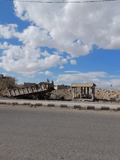 The image depicts a static daytime scene in Al-Qadam Municipality, Syria, characterized by significant structural damage and debris. In the foreground, a gray asphalt road occupies the lower portion of the frame, bordered on the right by a grey and black curb. Beyond the curb, a strip of dirt and minor debris separates the road from a large expanse of rubble piles that dominate the midground.

To the left of the midground, a large, heavily damaged, and rusty metal structure, possibly a section of a truck bed or industrial container, rests tilted upwards on the rubble, indicating extensive damage. To the right of the midground, situated atop or adjacent to the rubble, is a small, intact stone or concrete structure with four columns supporting a flat roof, resembling a small open shrine or kiosk. This structure appears weathered but not demolished.

In the background, further beyond the main rubble piles, skeletal remains of damaged buildings or structures are visible. The distant horizon to the right shows faint outlines of what appear to be hills or mountains. A few bare tree branches are visible on the far left edge of the image. The sky above is bright blue with numerous scattered white cumulus clouds. No individuals are visible, and no actions or interactions are depicted. Overhead, thin black lines, consistent with power lines, traverse the upper portion of the sky.