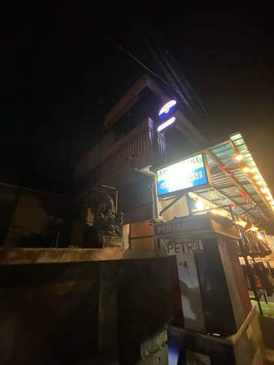 The image captures a nighttime scene on a street in Kuta Selatan, Indonesia, featuring commercial establishments. The primary focus is on a building with a prominent sign that reads "KING SMART GUEST HOUSE." Below this, another sign indicates "PERTAMINI PETROL," suggesting a gas station or fuel service. The structures appear to be made of concrete and wood, with a corrugated metal roof illuminated by strings of lights, creating a festive atmosphere. A dark, possibly stone, statue resembling an elephant or a mythical creature is visible to the left of the building, adding a cultural element to the scene. The overall environment is dimly lit, typical of a street at night, with no people clearly visible in the frame. Power lines are evident crisscrossing the dark sky above.