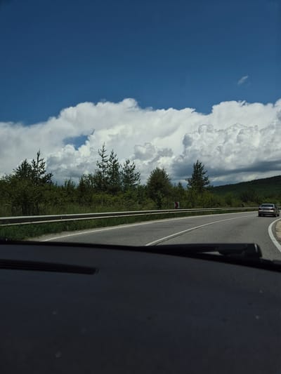 The image captures a first-person perspective from inside a vehicle traveling on a paved asphalt road in Ponor, Bulgaria, during daytime under a partly cloudy sky. The upper portion of the frame features a bright blue sky interspersed with large, white cumulus clouds, with some darker clouds visible towards the right. A dark green, tree-covered hill or mountain range is present on the far right horizon.
The midground displays a two-lane road curving to the right, marked with a white dashed center line and a solid white line along its right edge. A silver-colored sedan, consistent with a Mercedes-Benz C-Class (W203 generation), is visible ahead on the road, moving away from the viewer. Along the left side of the road, a metal guardrail separates the roadway from an embankment covered in dense green vegetation, including various trees resembling pines and numerous shrubs. Two red and white chevron signs, indicating a sharp right curve, are positioned on the right side of the road behind the guardrail.
The foreground is occupied by the dark-colored dashboard and the base of the windshield of the vehicle from which the photograph was taken. A black windshield wiper arm is partially visible on the right side of the dashboard. No individuals are explicitly visible in the scene. The overall lighting suggests clear daylight conditions.