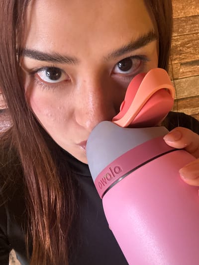A close-up, waist-up shot captures a person's face, with their left eye prominently in view. The individual, with long, dark brown hair, is looking directly at the camera. They are holding a pink and coral-colored water bottle with the brand name "Owala" visible on the gray rim. The background features a textured, stone-like wall in a light brown hue, suggesting an indoor environment. The lighting is bright, highlighting the subject's skin and the glossy finish of the water bottle. There are no visible landmarks or context that would indicate a specific location within San Andrés Cholula, Mexico.