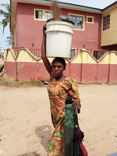 In Zango-ndaji, Nigeria, a woman stands on a dirt ground. She is carrying a white bucket filled with brooms on her head. She wears a long-sleeved, patterned dress. In her hand, she holds a rolled mat and an umbrella. Behind her is a building painted in shades of red and yellow, enclosed by a fence. A palm tree is visible in the upper left corner of the image.