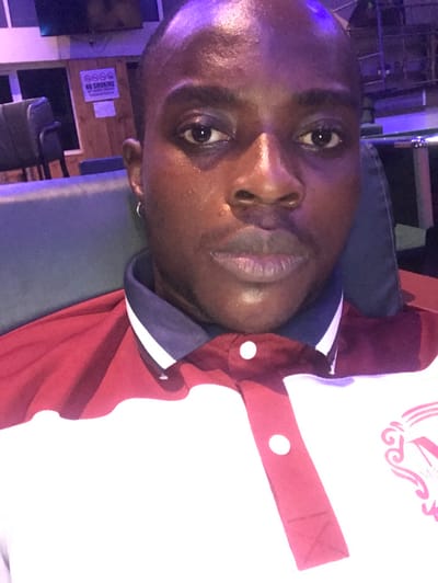 A male individual with dark skin, short dark hair, and a neutral expression is captured in a close-up, front-facing image, looking directly into the camera. He is wearing a short-sleeved polo shirt with a white main body, a dark blue collar, and dark red panels on the shoulders and button placket. Two white buttons are visible on the placket. A small silver hoop earring is present in his left ear. A partial pink graphic or logo is visible on the lower right side of his shirt.

The setting is an indoor, dimly lit environment, possibly a lounge or similar commercial establishment, characterized by a prominent purple and blue ambient lighting scheme. In the background, dark upholstered seating is visible, along with a segment of a wooden wall. A white sign with red text, featuring a 'NO SMOKING' emblem and additional unreadable text below, is affixed to the wooden wall. A dark rectangular object, appearing to be a monitor or television, is mounted high on the wall in the upper left background. The location is stated as Lekki, Nigeria.