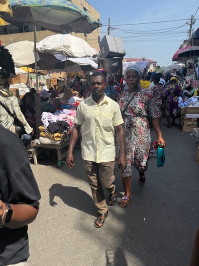 The image depicts a crowded outdoor market in Accra, Ghana, under bright daylight. In the immediate foreground, to the left, the left shoulder and arm of an individual wearing a black t-shirt and a gold-colored smartwatch are visible. Centered in the mid-ground, a dark-skinned adult male with short dark hair and stubble walks forward, looking directly at the camera. He wears a light yellow short-sleeved collared shirt, brown trousers, and black sandals. To his right and slightly behind him, a dark-skinned adult female also walks forward, looking towards the camera. She wears a light-colored headscarf, a floral-patterned knee-length dress, and open-toed sandals, carrying a small turquoise-green handbag in her left hand.

The background reveals a dense market environment with numerous people and vendor stalls. Multiple large, light-colored umbrellas and tarpaulins provide shade over various makeshift stands, which display goods including piles of clothing, textiles, and stacked cardboard boxes. A wooden table holding white and black garments is visible to the left. In the mid-background, individuals are seen navigating the crowd; one person carries a large silver rectangular container on their head, and another carries a smaller silver bowl. A multi-story light-colored building is partially visible in the upper-left background. Overhead utility wires crisscross the sky. The ground is paved with light-colored blocks or pavers, and strong sunlight casts distinct shadows, particularly from the man in the foreground.