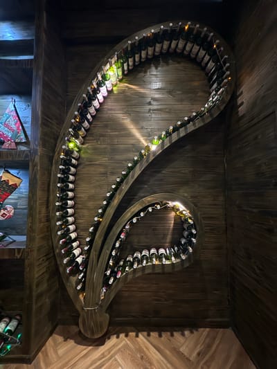 The image shows an elaborately designed wooden wine rack that resembles a spiral or a paisley shape, mounted on a wooden-paneled wall. The rack holds numerous wine bottles, many of which are lit by small, string lights that are intertwined with the bottles. The overall aesthetic is rustic and dimly lit, with warm tones from the wood and the string lights. The wine rack itself is a prominent art installation. On the left side of the image, there are shelves with decorative items, including what appear to be Christmas stockings and a fabric wall hanging. The flooring is also made of wood, laid in a parquet pattern. This scene is located in a private establishment or residence, likely a cellar or a dedicated display area, in Da Lat, Vietnam, known for its cool climate and French colonial architecture, which often features wooden elements and intimate settings suitable for wine appreciation. No people are visible in the image.