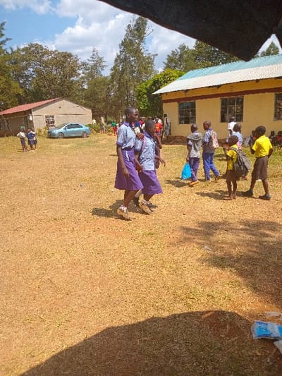 The image shows a scene outside a building in Malava, Kenya, located at coordinates 0.53 latitude and 34.811 longitude. The building appears to be a school, with students in uniform gathered in the yard. There is a group of four students in the foreground, two wearing purple skirts and two wearing blue shirts. The students are walking on a grassy field. They are interacting with each other. The background shows the building, and a car parked nearby, with a tree partially visible. The sun is out, and the ground appears dry and dusty. The building is partially visible and has a yellow roof, blue trim, and white walls. The building appears to have a school logo. The scene is taken from a slightly elevated perspective and appears to be captured by a person moving forward while holding the camera.  The scene is bright and well-lit, and there is a clear blue sky in the background.