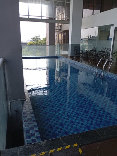 The image depicts an indoor swimming pool area, likely a part of a hotel or fitness center, located in South Jakarta, Indonesia. The pool itself is rectangular, tiled with a pattern of blue and darker blue squares. The water surface reflects the bright light from large windows that overlook an outdoor area with trees and buildings. Adjacent to the pool, there is a seating area with several dark-colored chairs and tables. A metal ladder is submerged in the pool, providing access. The surrounding environment includes modern architecture with glass walls and pillars. The scene is clean and appears well-maintained, suggesting a leisure or recreational facility. There are no people visible in the image.