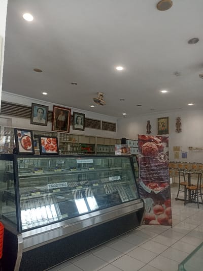 The scene is the interior of a bakery located at coordinates -7.428, 109.247 in Purwokerto, Indonesia. The space is brightly lit with fluorescent lights and features a glass display case filled with baked goods. The display case has labels identifying the goods as "Traditional" and "Modern." Two framed pictures of individuals are visible on the wall to the left of the display case. A large poster of a bakery item is located to the right of the display case. There are white tiled floors and some shelving units in the background. There are three tables and chairs to the right of the display case, which appear to be empty.