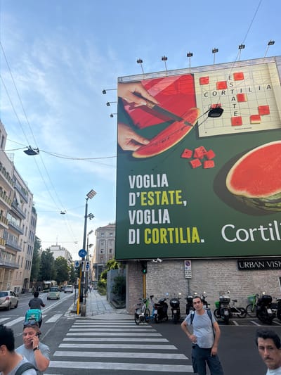This vertical-format photograph captures a street scene in Milan, Italy, under a clear, blue sky with scattered clouds, suggesting a sunny day. Dominating the upper portion of the image is a large billboard advertising "Cortilia," an Italian e-commerce platform for food. The billboard features a close-up of a hand slicing a watermelon, surrounded by a crossword puzzle with Italian words related to summer and Cortilia. The text "VOGLIA D'ESTATE, VOGLIA DI CORTILIA" (Desire for Summer, Desire for Cortilia) is prominently displayed.
Below the billboard, a street unfolds with a pedestrian crossing and parked motorbikes. Several people are visible. In the foreground on the right, a man in a light grey t-shirt and dark pants is standing near the pedestrian crossing, looking towards the camera and holding a phone to his ear. Another person, partially visible, is to his right. Further down the street, a person with a teal backpack is walking away from the camera. The urban setting includes multi-story buildings lining the street, some with visible windows and balconies. Traffic lights and street signs, including a parking sign and a bicycle lane sign, are also present. The overall impression is a typical bustling city day with commercial advertising and everyday life unfolding.