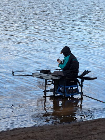 A man is fishing in a body of water. The man is seated in a blue fishing chair, wearing a black and teal jacket with the word "DREAM" visible on the back. He is hunched over, holding an object in his hands and looking down at it. A fishing rod is propped up behind the chair. A net is hanging from the side of the chair. The water is rippled, and the shoreline is visible on the right. The city of Chaykovsky, Russia, is not discernible from this image.