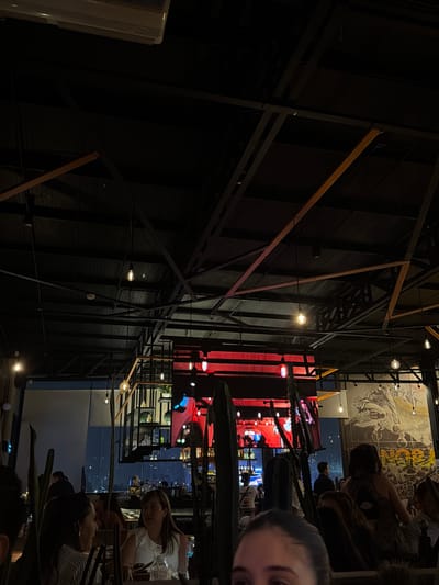 The image shows the interior of a restaurant or bar in Puebla, Mexico, based on the "MADISON" sign visible. Patrons are seated at tables, and a TV screen shows city lights. The interior features exposed structural elements of black metal and brown wood, with decorative lighting. A mural or large graphic is seen on an adjacent wall. A person's face is visible in the lower right corner of the image.