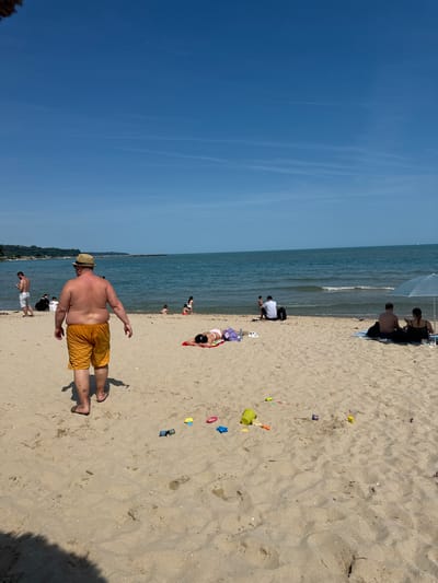 This image captures a busy sandy beach on a clear, sunny day in Varna, Bulgaria. The sky is a bright, unblemished blue with faint, wispy contrails. The blue-green sea extends to the horizon, featuring small waves breaking near the shore, with a distant sailboat visible on the right.

In the foreground, numerous colorful plastic beach toys, including buckets, shovels, and molds, are scattered on the light-colored sand. Dominating the left midground, a large-built, shirtless man wearing a straw hat and bright orange swim shorts, with flip-flops, walks away from the viewer towards the water, casting a long shadow. To his left, another man, shirtless and in white shorts, stands looking at his mobile phone. Further along the beach, closer to the water's edge, a group of individuals are seated on the sand, while a child is seen playing in the shallow waves with an adult nearby.

On the right side of the midground, a woman in a pink bikini top and purple bottoms is lying prone on a red towel, sunbathing. To her right, a light blue beach umbrella shades a couple seated on a striped towel; the man is shirtless, and the woman wears a dark top. In the background, a treeline runs along the coast, leading to a visible pier or jetty structure extending into the sea on the left. The bright sun casts distinct shadows across the sandy expanse, indicating direct overhead light.
